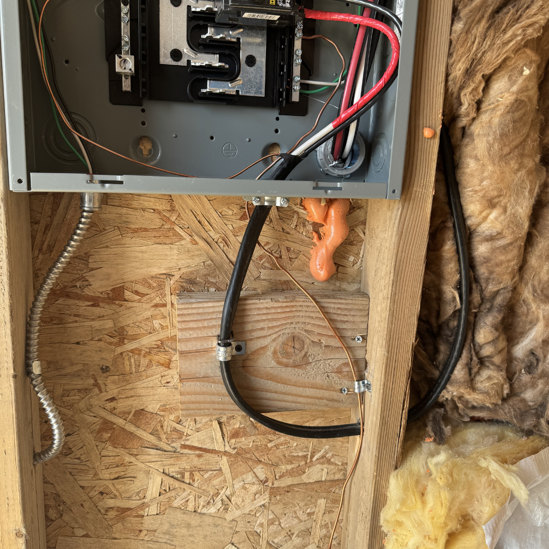 Electrical wiring inside an electrical panel mounted on a wooden board, with wires and a circuit breaker, next to insulation and some foam.