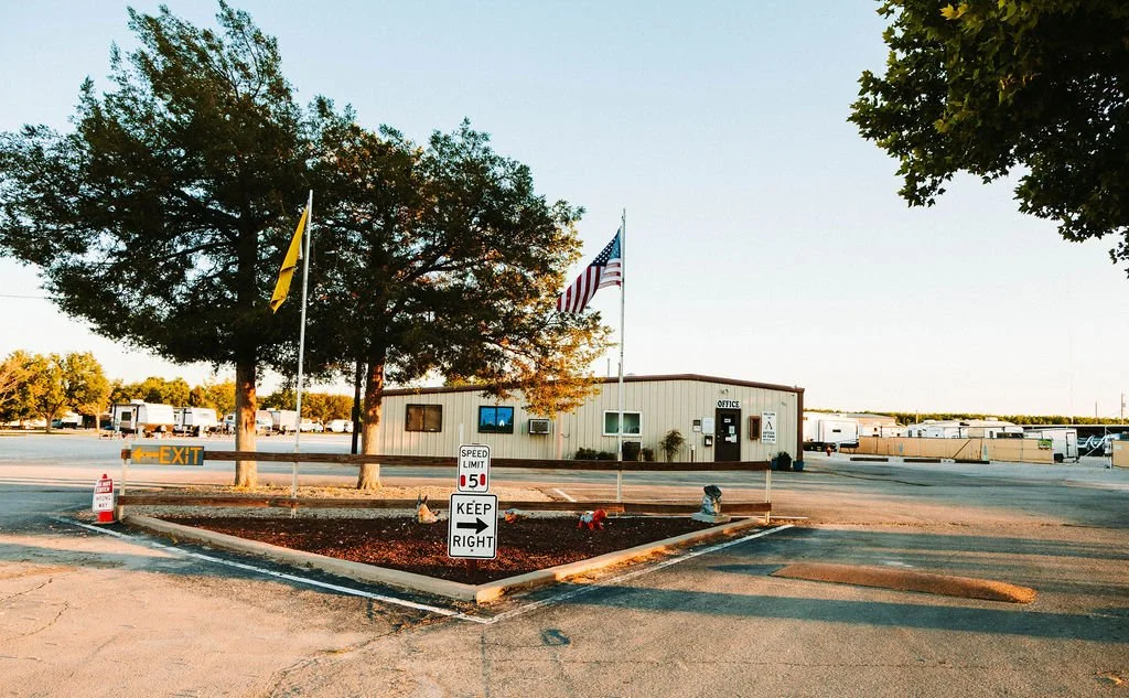 A parking lot with trees, two flags (American flag and yellow flag), a small office building, and several RVs or trailers in the background.