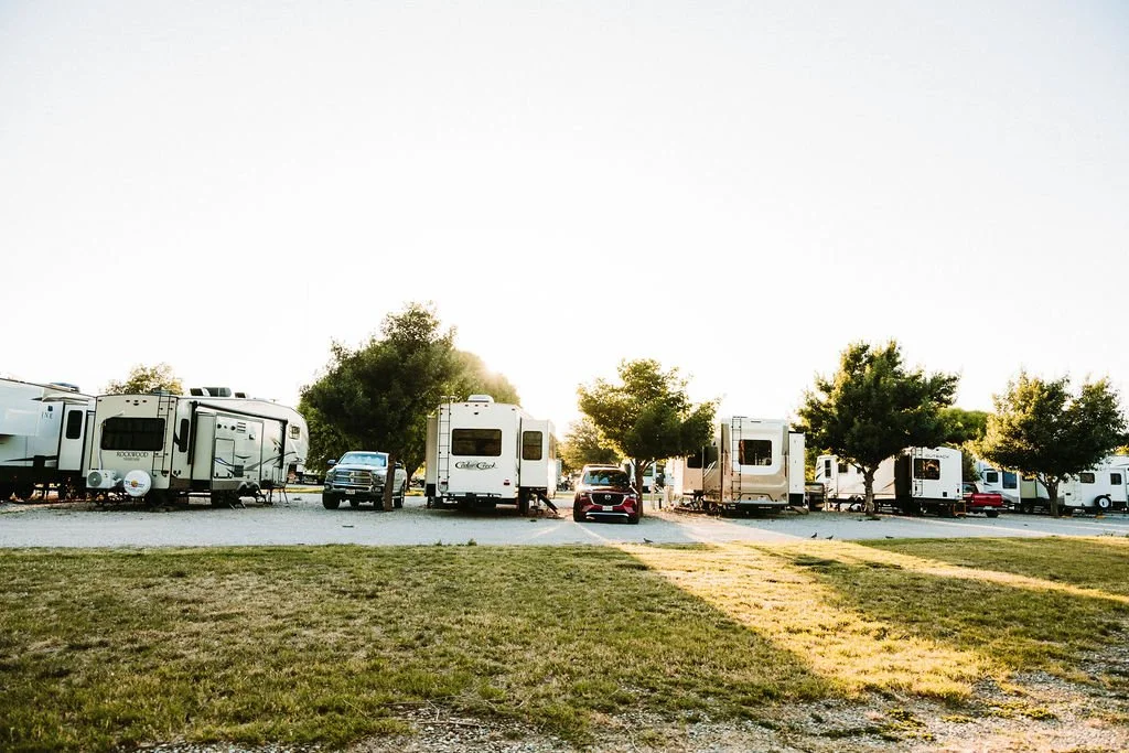 A row of parked RVs and cars in a campground with trees and grassy area, during sunset.