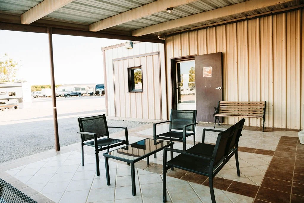 Outdoor patio area with black metal chairs and a glass-top table, attached to a building with vertical metal siding and a covered roof, in a parking lot.