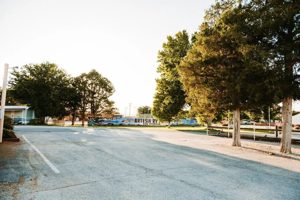 Empty parking lot with trees and a mural in the background that reads 'Artesia RV and Storage' during late afternoon or early evening.