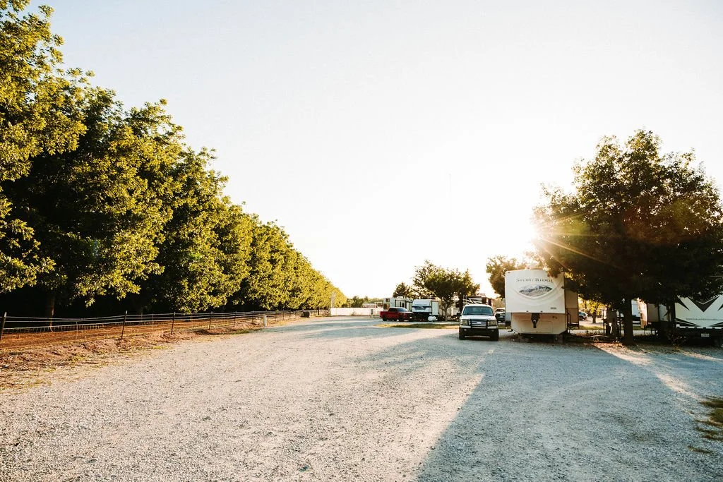A gravel parking lot with trees on the left and right, utility trucks, cars, and RVs parked, with a sunset in the background and sunlight lens flare.