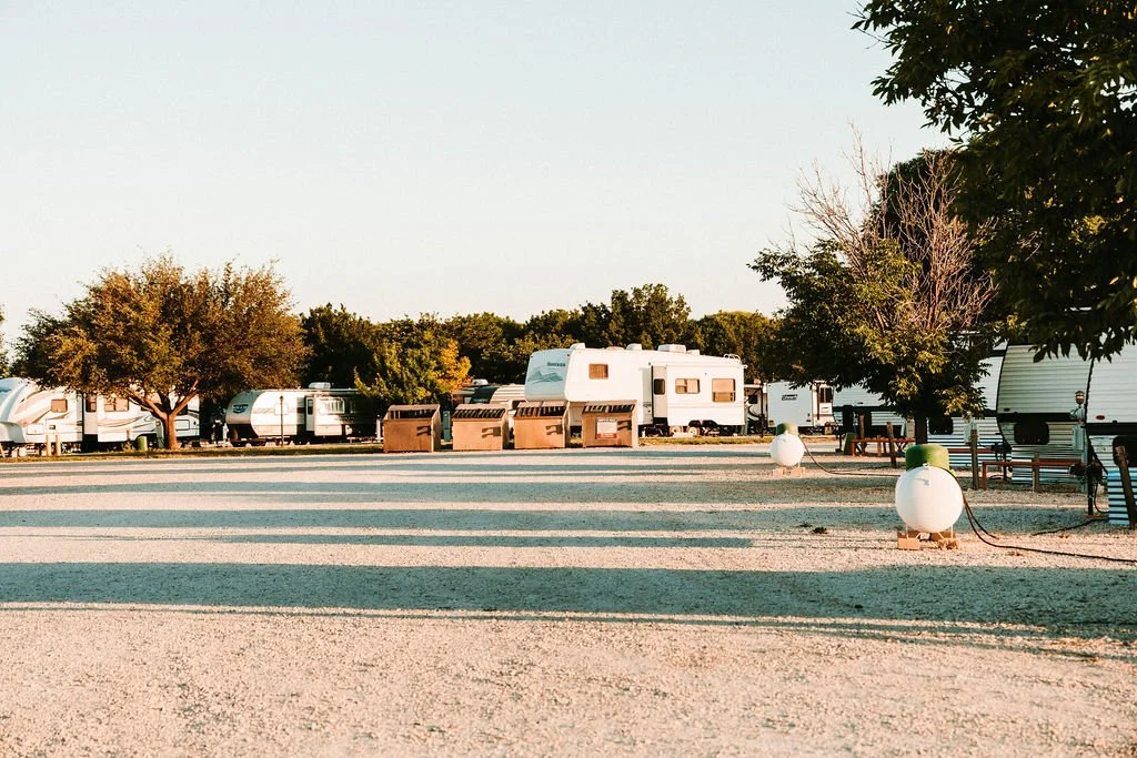A campground with multiple RVs and trailers parked among trees, with benches, trash cans, and propane tanks visible in the area.