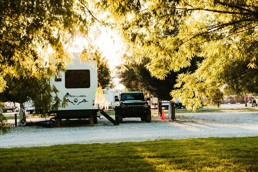 A black vehicle parked next to a white camper trailer with a sticker that reads 'Dock Label', surrounded by trees at sunset.