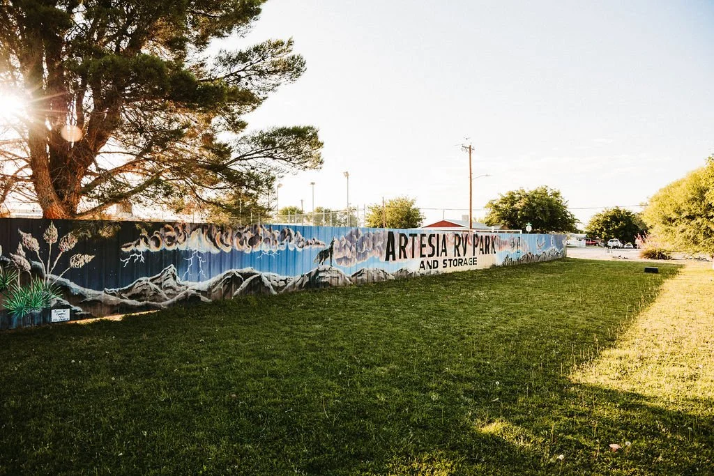 A mural on a fence reads 'Artesia RV Park and Storage,' with mountains and sky painted on it, in a grassy area with trees and sunlight.