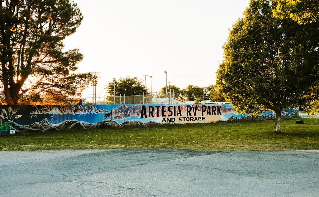 A mural on a wall at Artesia RV Park and Storage, with trees and power lines in the background, during sunset.