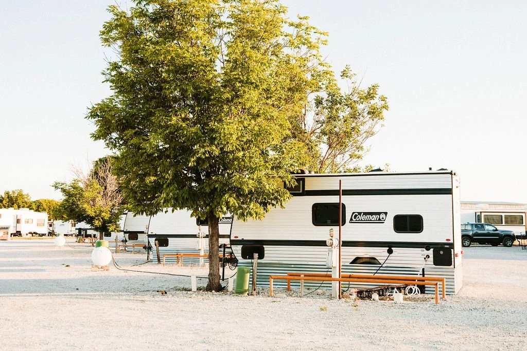 A large white Coleman travel trailer parked in an RV lot with a tree in front, surrounded by benches and outdoor equipment, and other RVs and vehicles in the background.