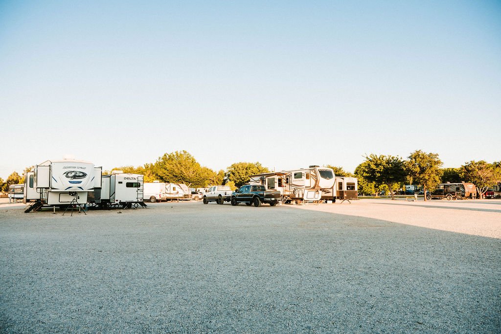 An RV parking lot with multiple RVs, trucks, and trees in the background under a clear sky.