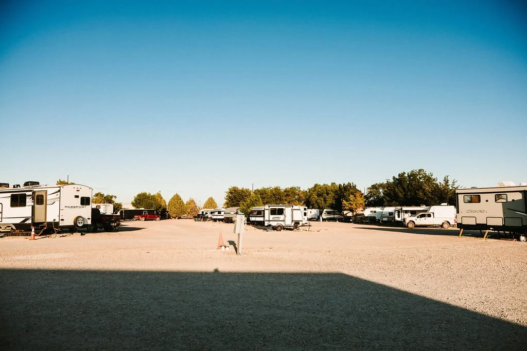 A gravel parking lot with multiple RVs and trailers parked, some trees and a clear blue sky in the background.