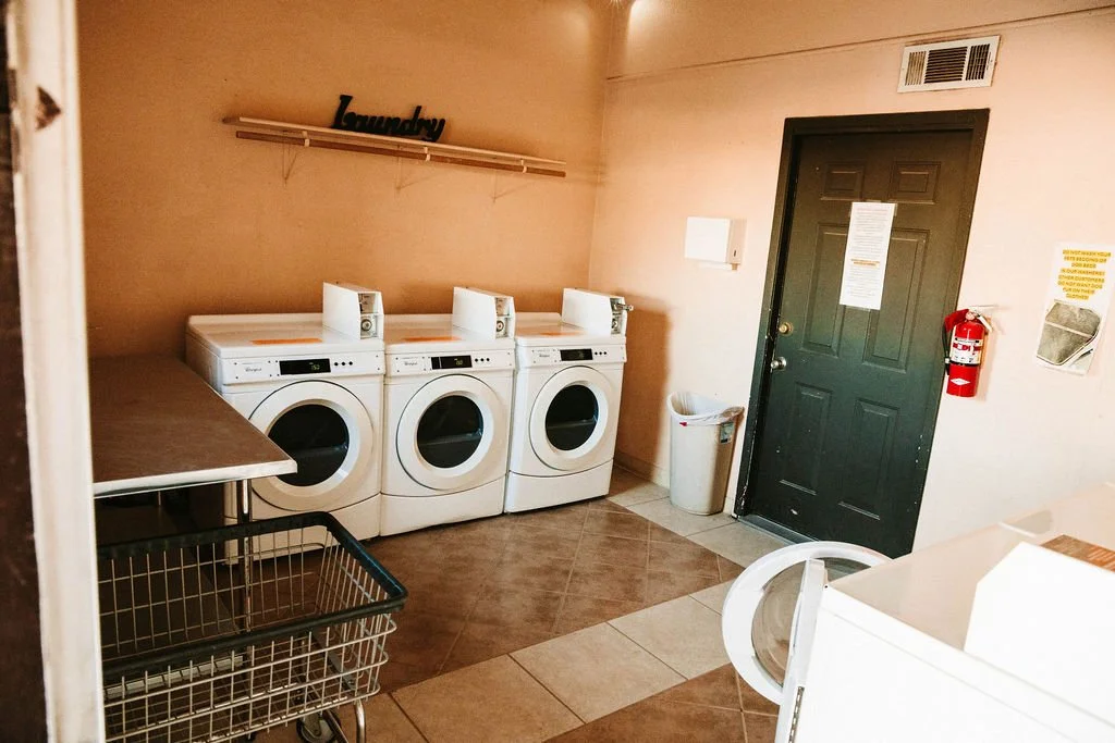 Laundry room with three washing machines, a laundry basket, a table, a trash can, a fire extinguisher, and a black door.