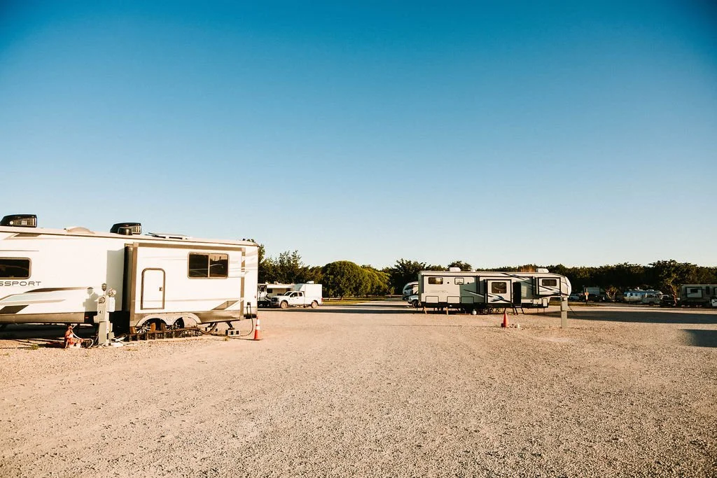 Rv trailers parked on a gravel lot with few cars and trees in the background and a clear blue sky.