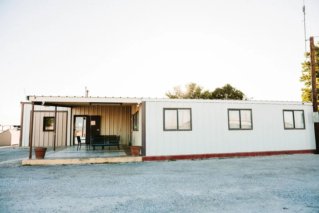 A small, single-story building with a covered front porch, two potted plants, three windows, and a black door, located on an unpaved gravel lot.