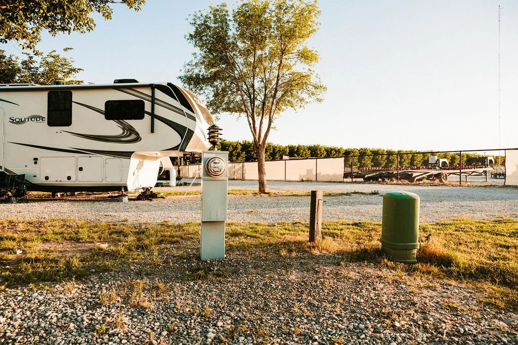 A recreational vehicle parked at a campsite with a large tree nearby, electrical hookup, and utility boxes, enclosed by a chain-link fence, with trailers in the background during sunset.