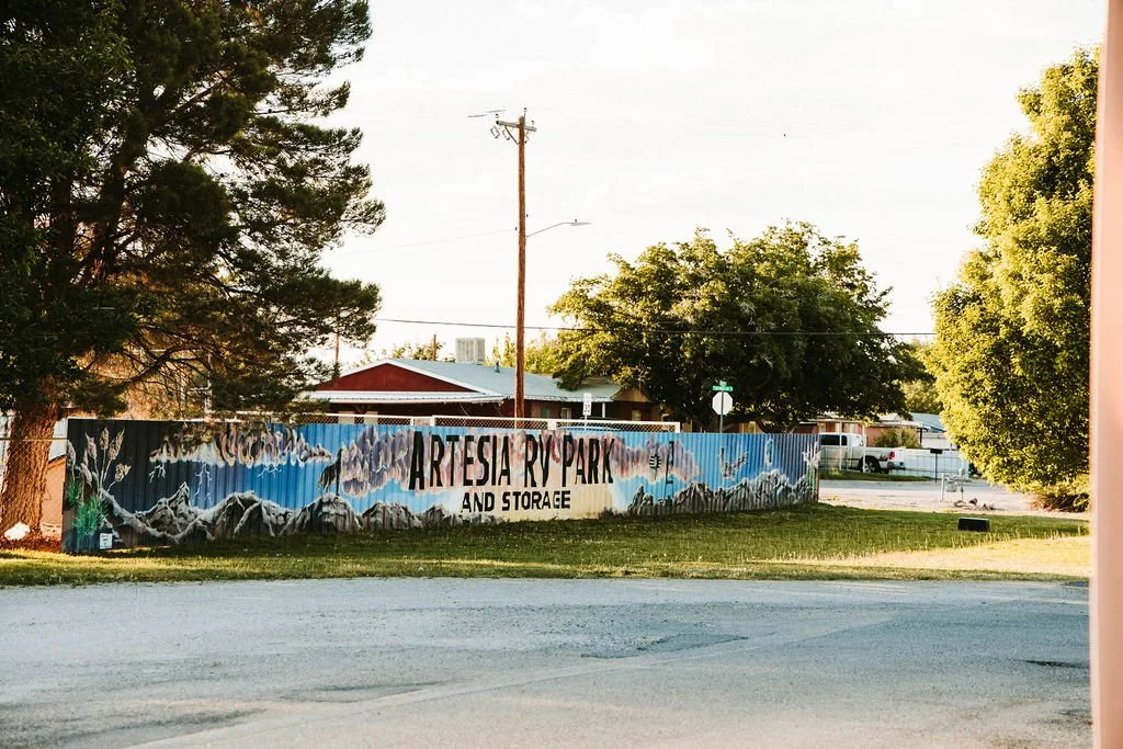 Painted sign reading Artesia RV Park and Storage with a mountain landscape, trees, and a parking lot in the background.