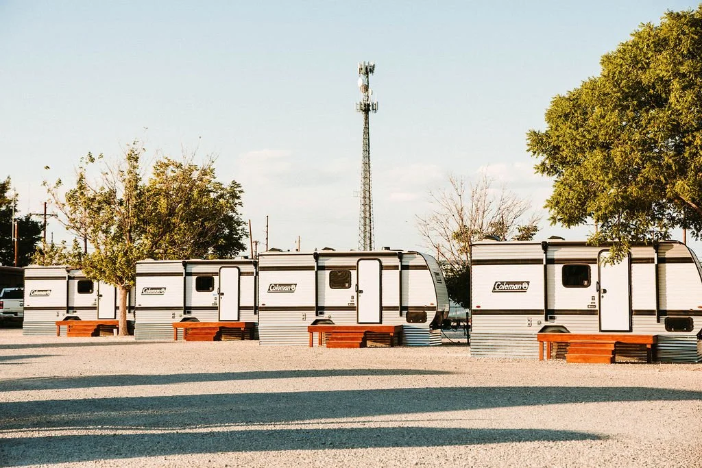 Three Coleman travel trailers parked in a lot with trees and benches in front, during daylight.