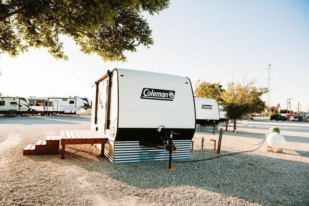A Coleman travel trailer parked on a gravel lot, with wooden steps leading up to the entrance, surrounded by trees and other RVs in the background, under a clear sky.