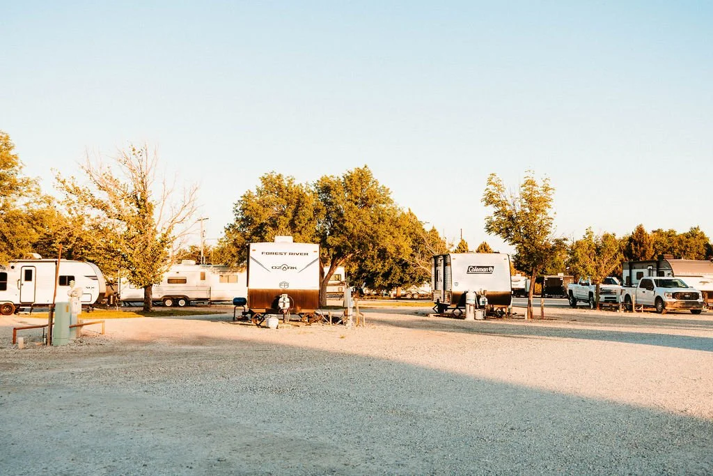 RV campground with travel trailers, trees, and parked trucks in a sunny outdoor setting.