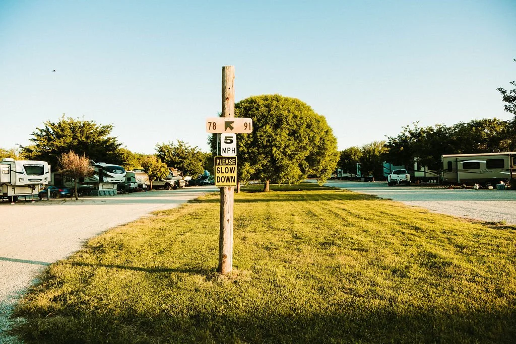 A grassy area with a utility pole and multiple signs, including a speed limit of 5 MPH and a reminder to slow down. In the background, there is an RV park with several parked RVs and trees, under a clear sky.