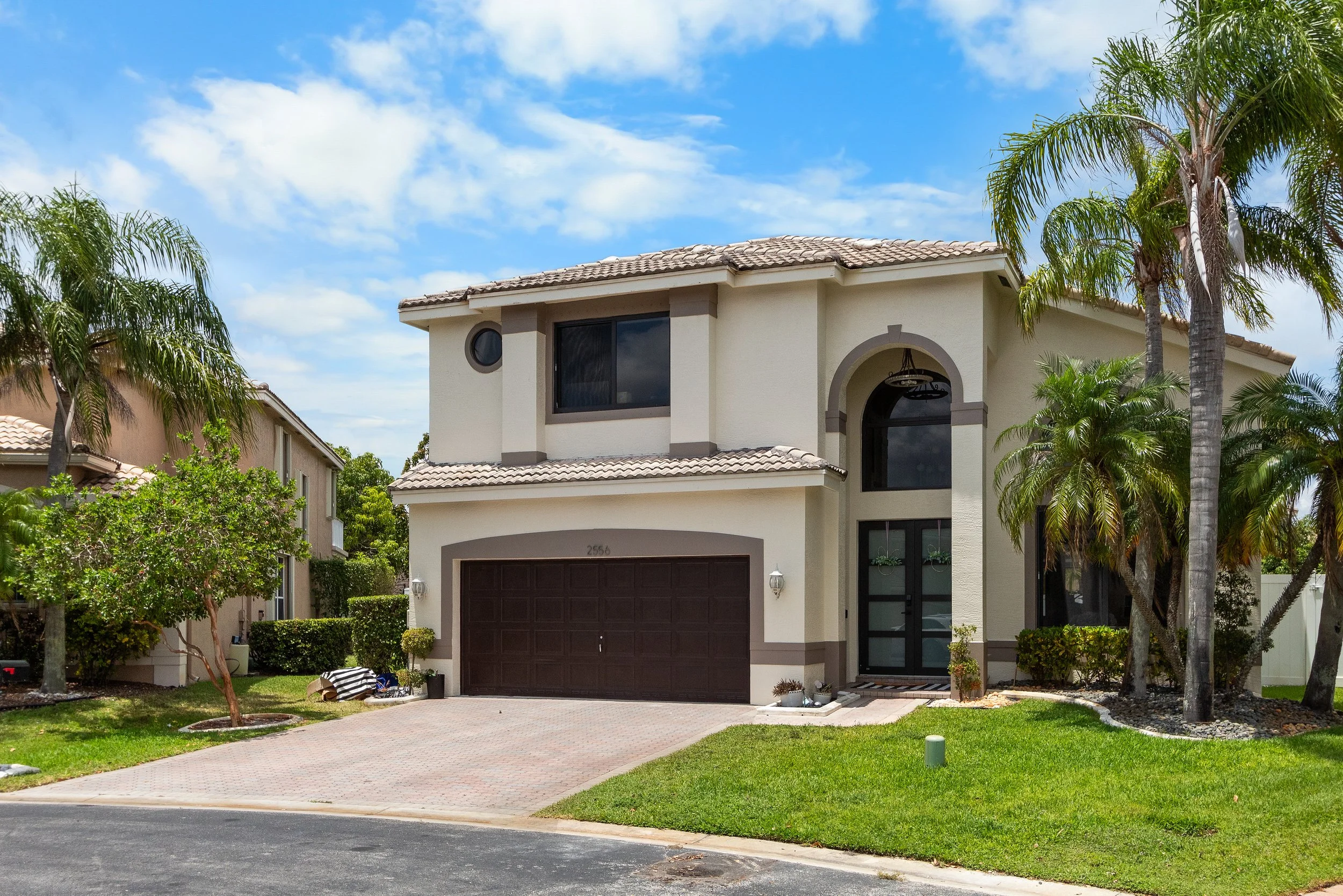 Front view of a two-story house with a beige exterior, dark brown garage door, and surrounding palm trees, under a partly cloudy sky.
