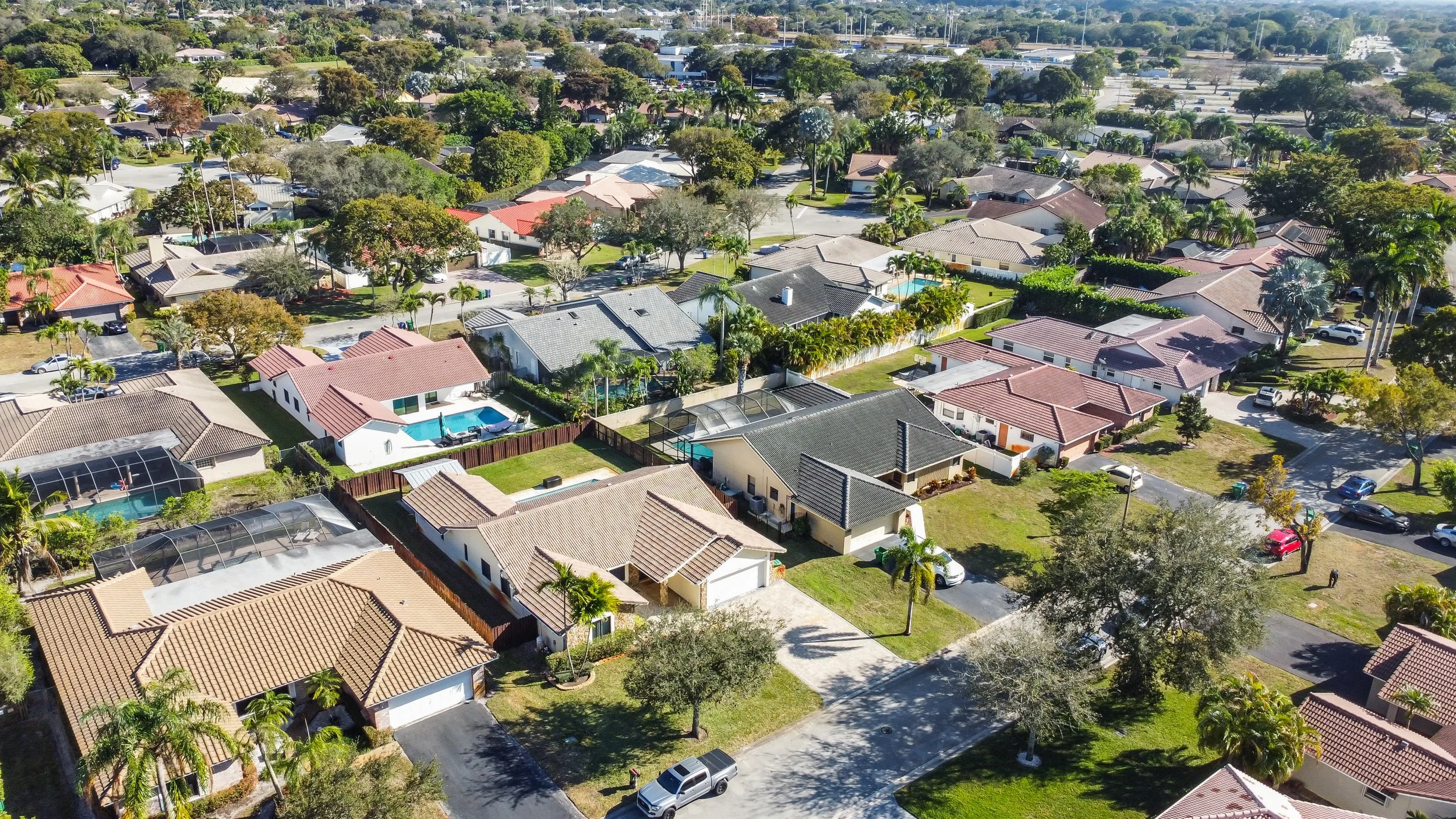 Aerial view of a suburban neighborhood with single-family homes, some with swimming pools, surrounded by trees and well-maintained lawns.