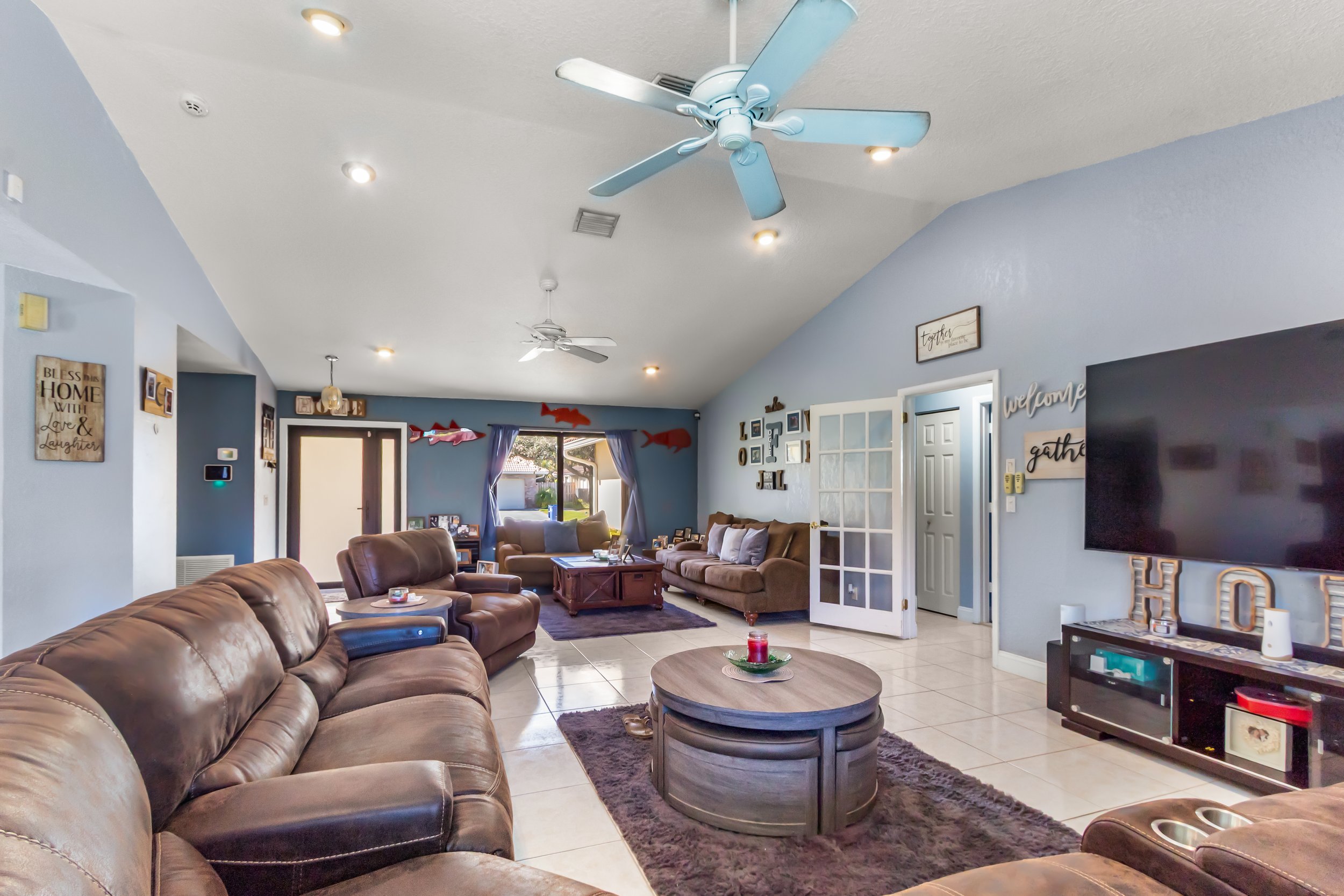 Living room with multiple brown leather sofas, a central round coffee table, a large flat-screen TV on the wall, ceiling fans, and a window with blue curtains, decorated with framed pictures, wall signs, and plush toys.
