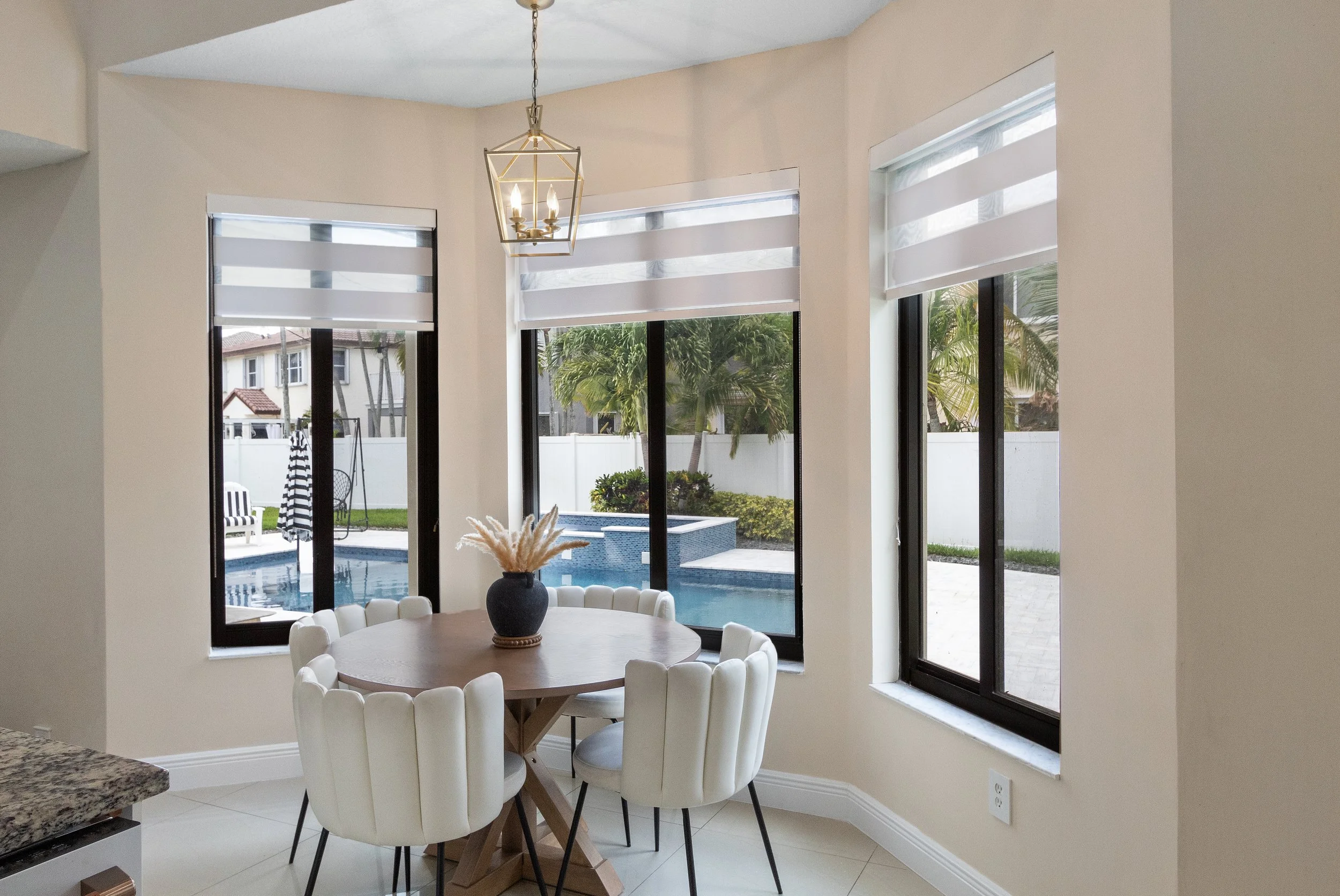 A dining area with a round wooden table surrounded by six white upholstered chairs, a black vase with dried pampas grass on the table, large windows showing a backyard with a pool, trees, and a white fence, and a gold geometric chandelier hanging from the ceiling.