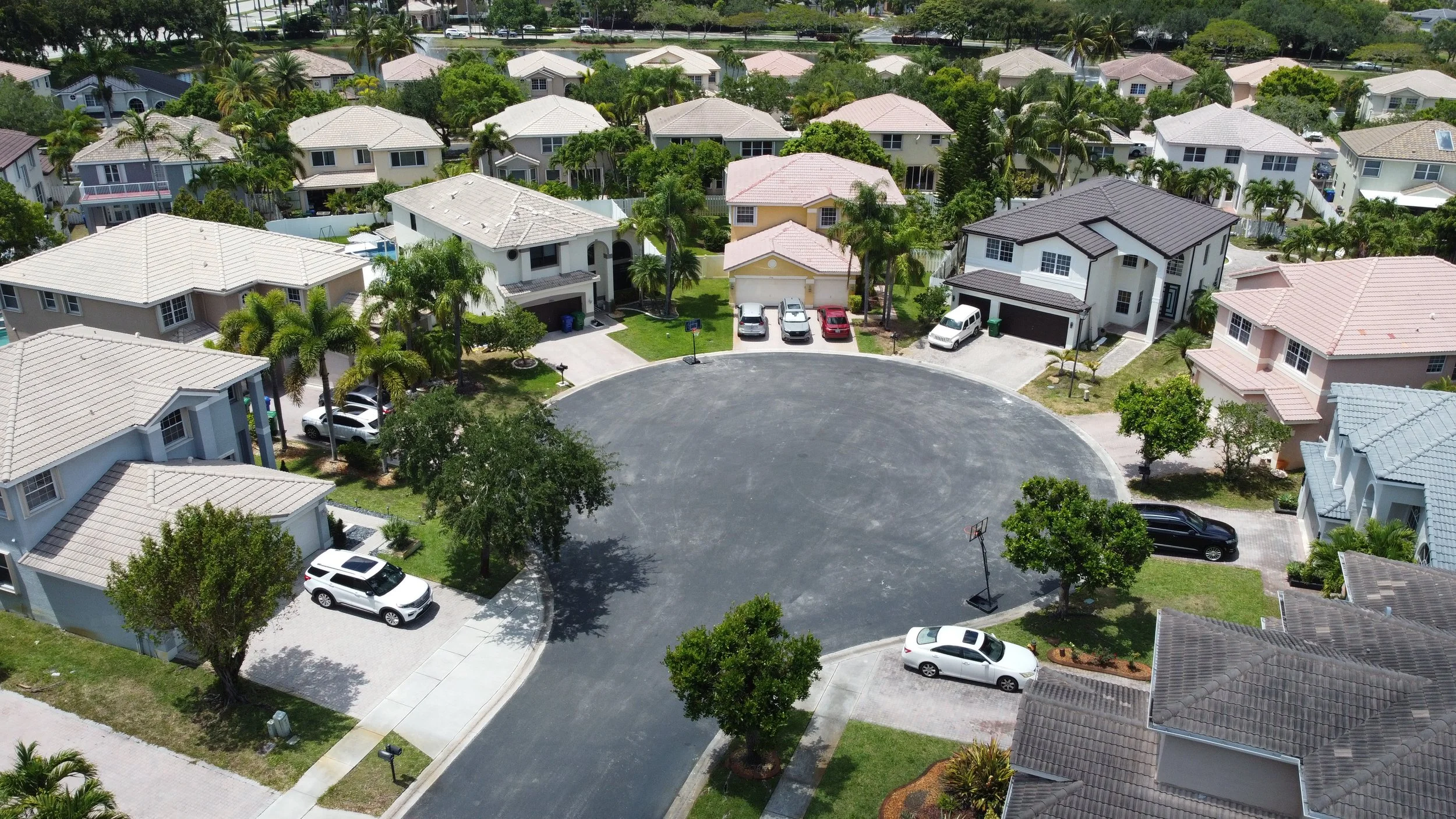 An aerial view of a suburban neighborhood with multiple houses, palm trees, parked cars, and a circular asphalt cul-de-sac.