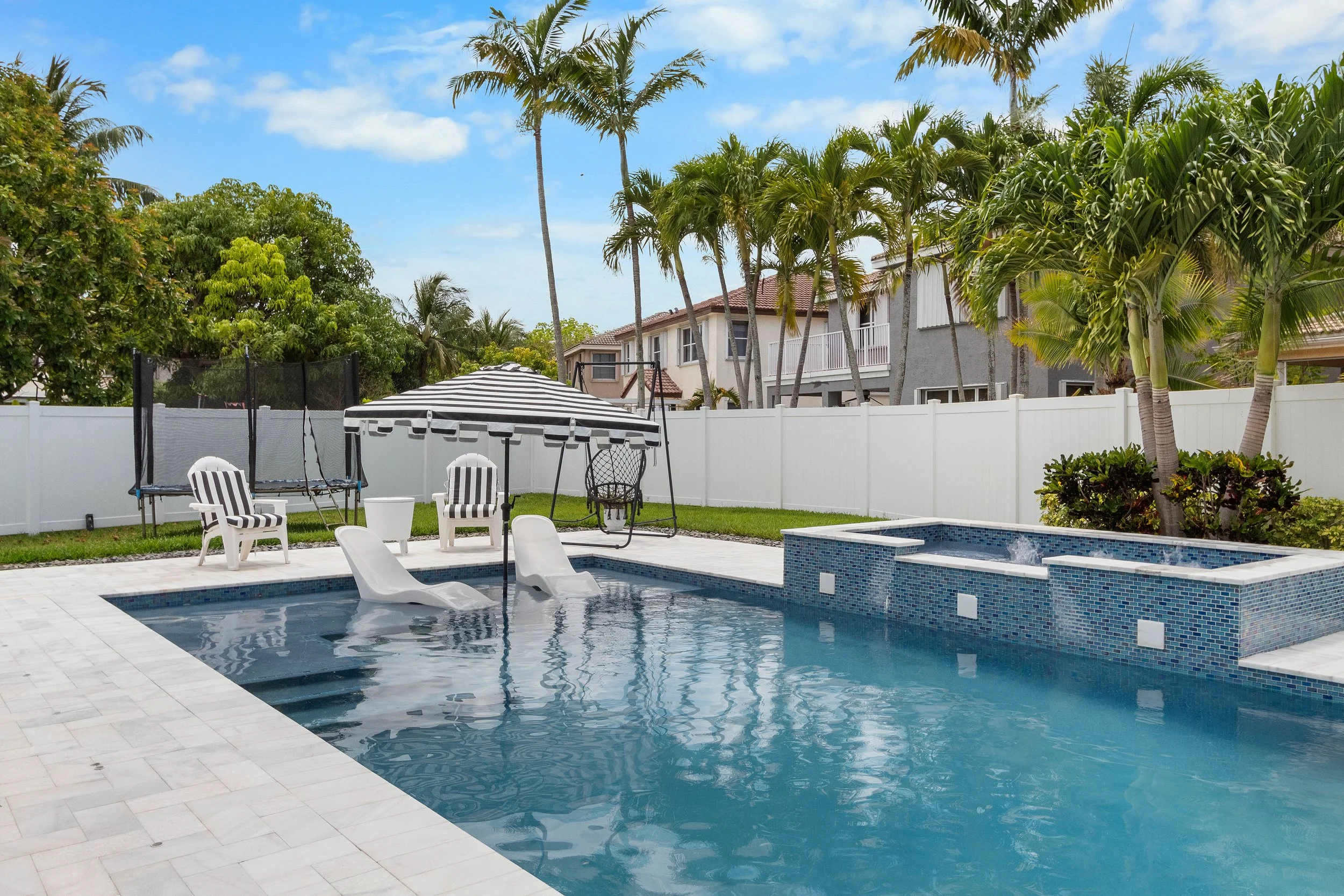 Backyard swimming pool with white tiled deck, two plastic slides, two striped lounge chairs, a patio umbrella, a small trampoline, and surrounding palm trees and residential houses.