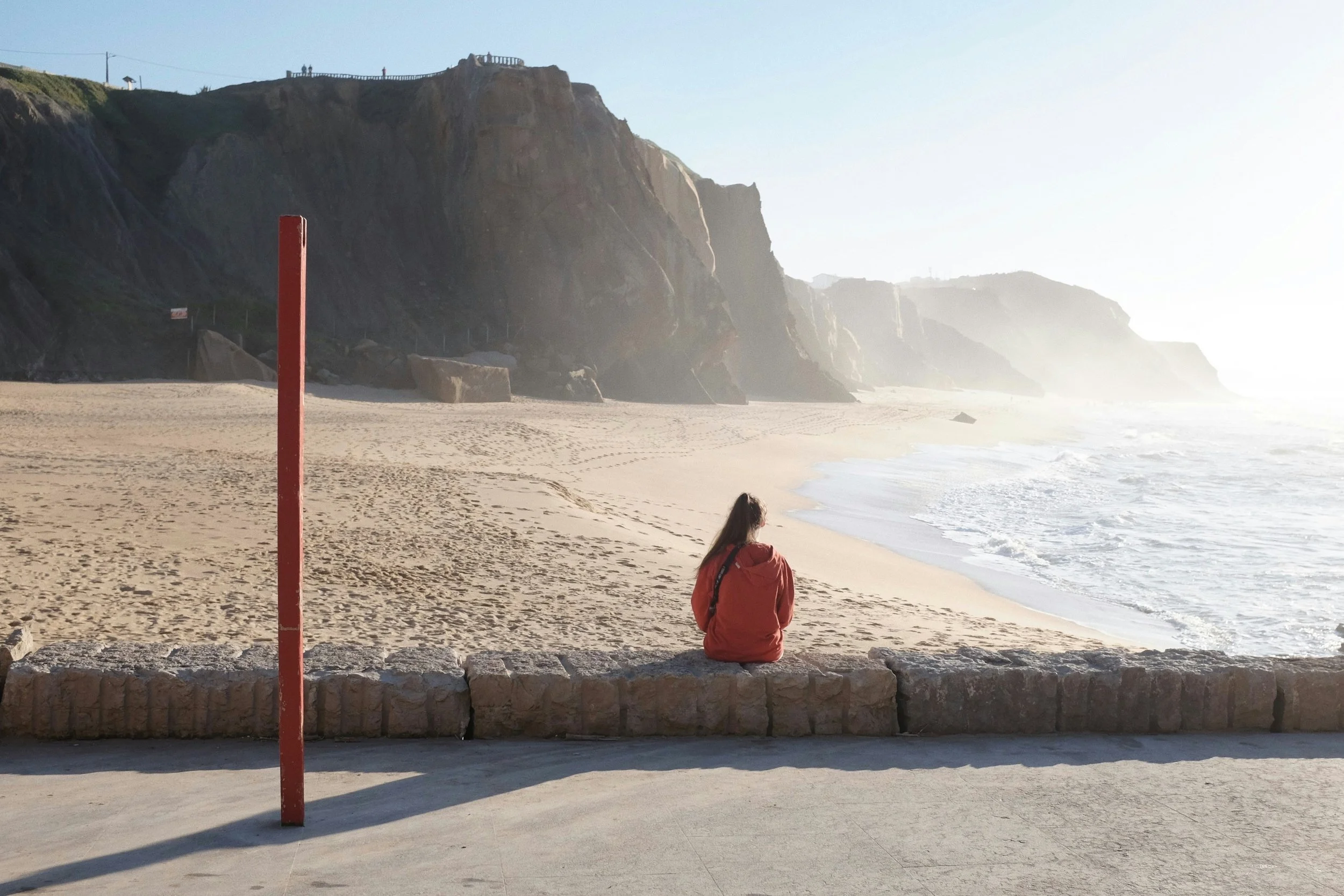 A person sitting on a stone wall at a beach, looking towards the ocean and cliffs in the distance