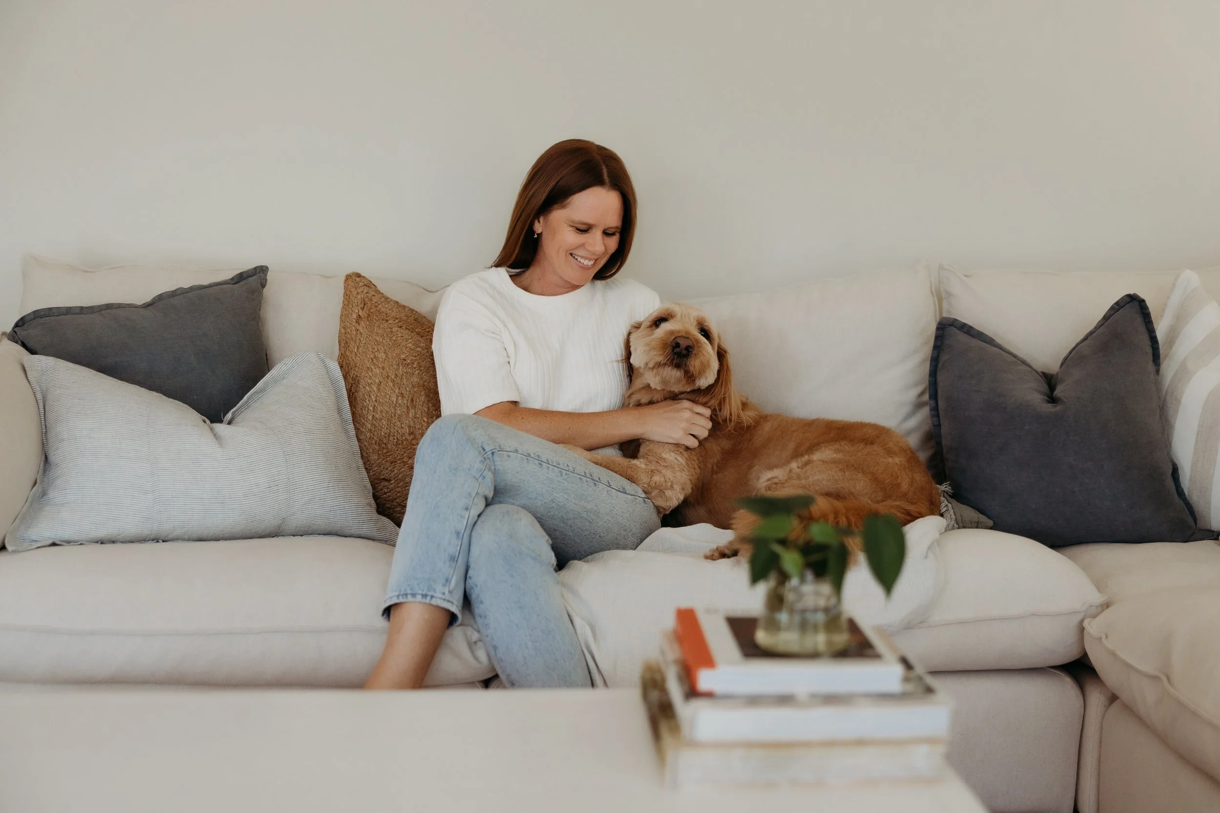 A woman sitting on a cream-colored sofa, smiling while hugging a large, relaxed brown dog with a scruffy coat around her.