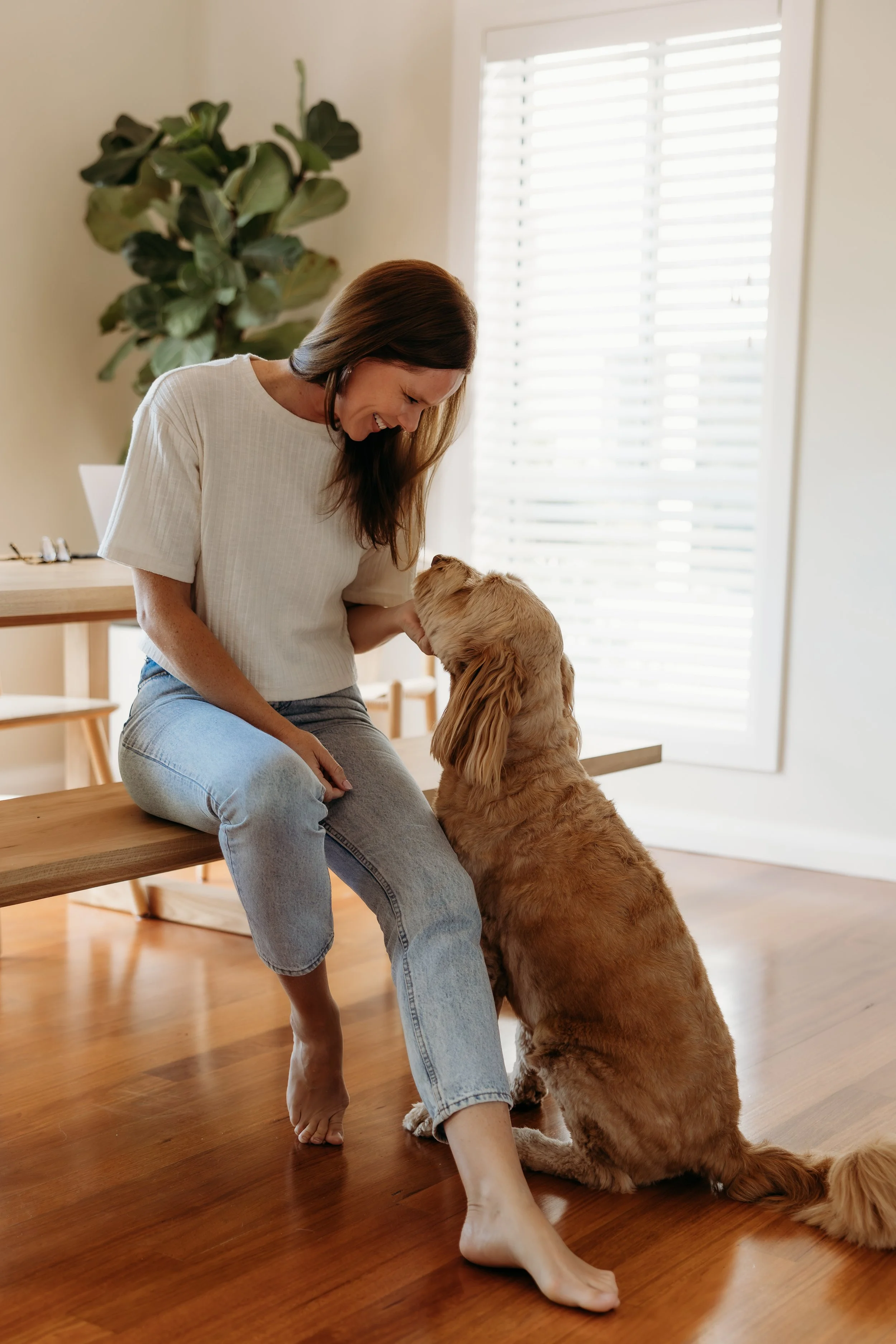 A woman sitting on a wooden bench smiling at a golden retriever dog that is sitting on the wooden floor and looking at her.