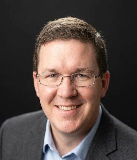 Headshot of Bill Dalbec, founder and CEO of Baseline Strategic Partners, smiling with glasses, wearing a dark blazer and light blue shirt, against a black background.