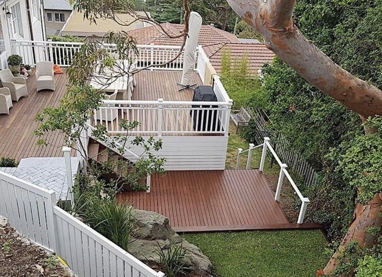 A multi-level backyard with wooden deck areas, outdoor seating, a barbecue, views of neighboring rooftops, and surrounding greenery including trees and bushes.