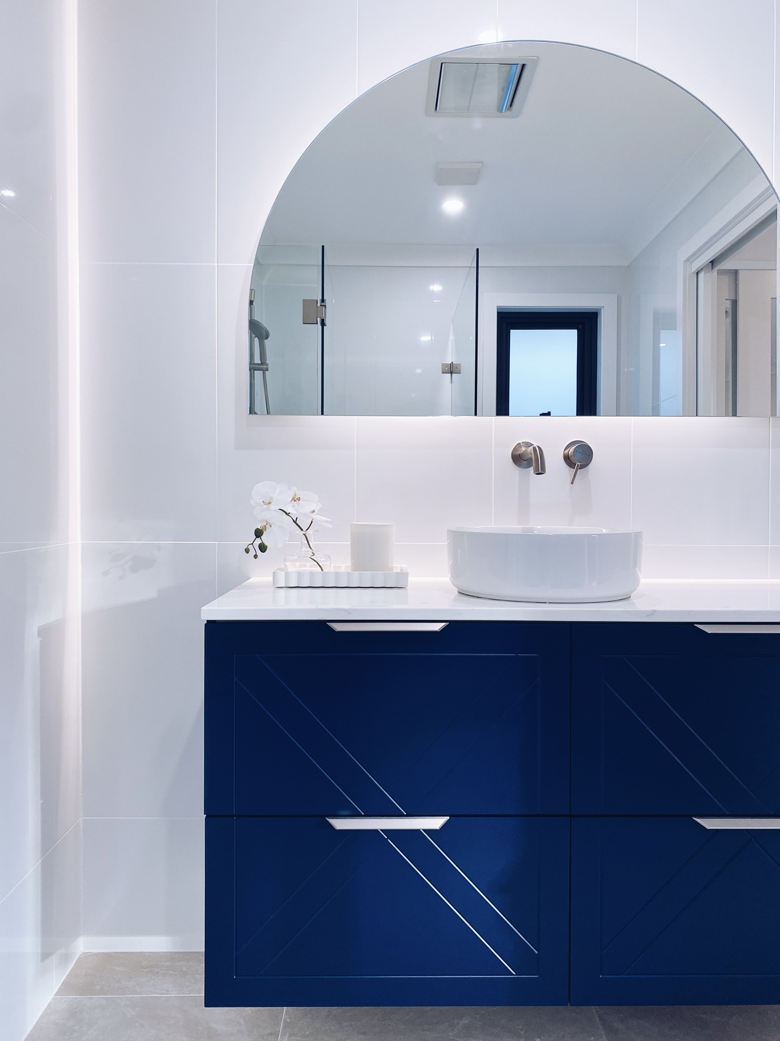 Modern bathroom with white walls, a large mirror, a blue cabinet vanity with silver handles, a white vessel sink, a wall-mounted faucet, a small orchid plant, and a shower with a glass door and a showerhead in the background.
