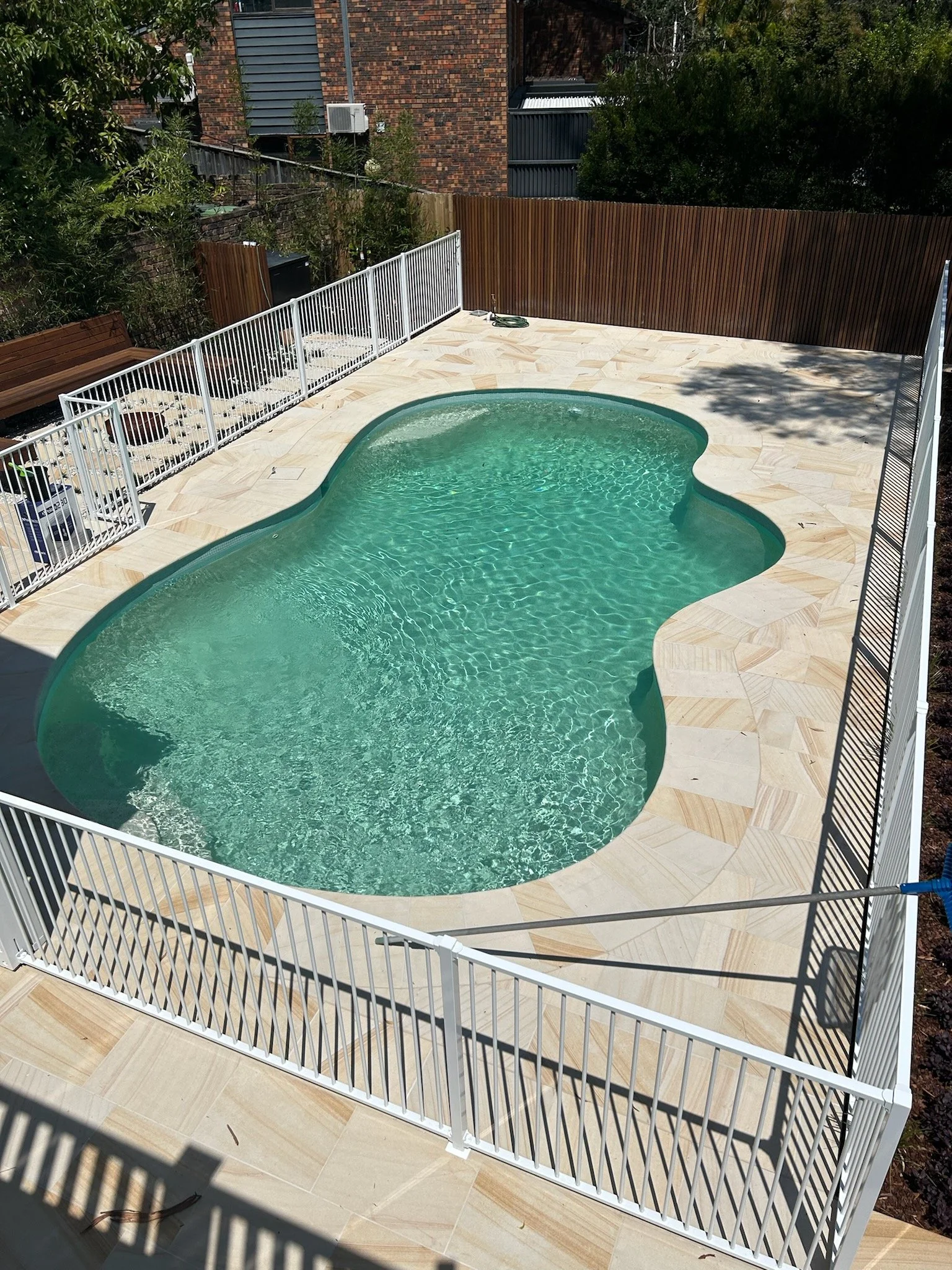 An outdoor backyard with a kidney-shaped swimming pool, surrounded by a beige tile deck and a white metal fence. There is a wooden bench on the left side, and a brown wooden privacy fence at the back. Some trees and bushes are visible around the yard