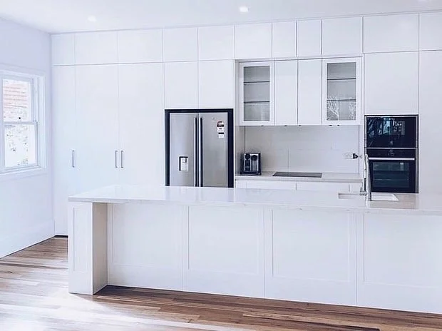 Modern white kitchen with a large island, stainless steel refrigerator, built-in oven, microwave, and cabinets with glass doors.