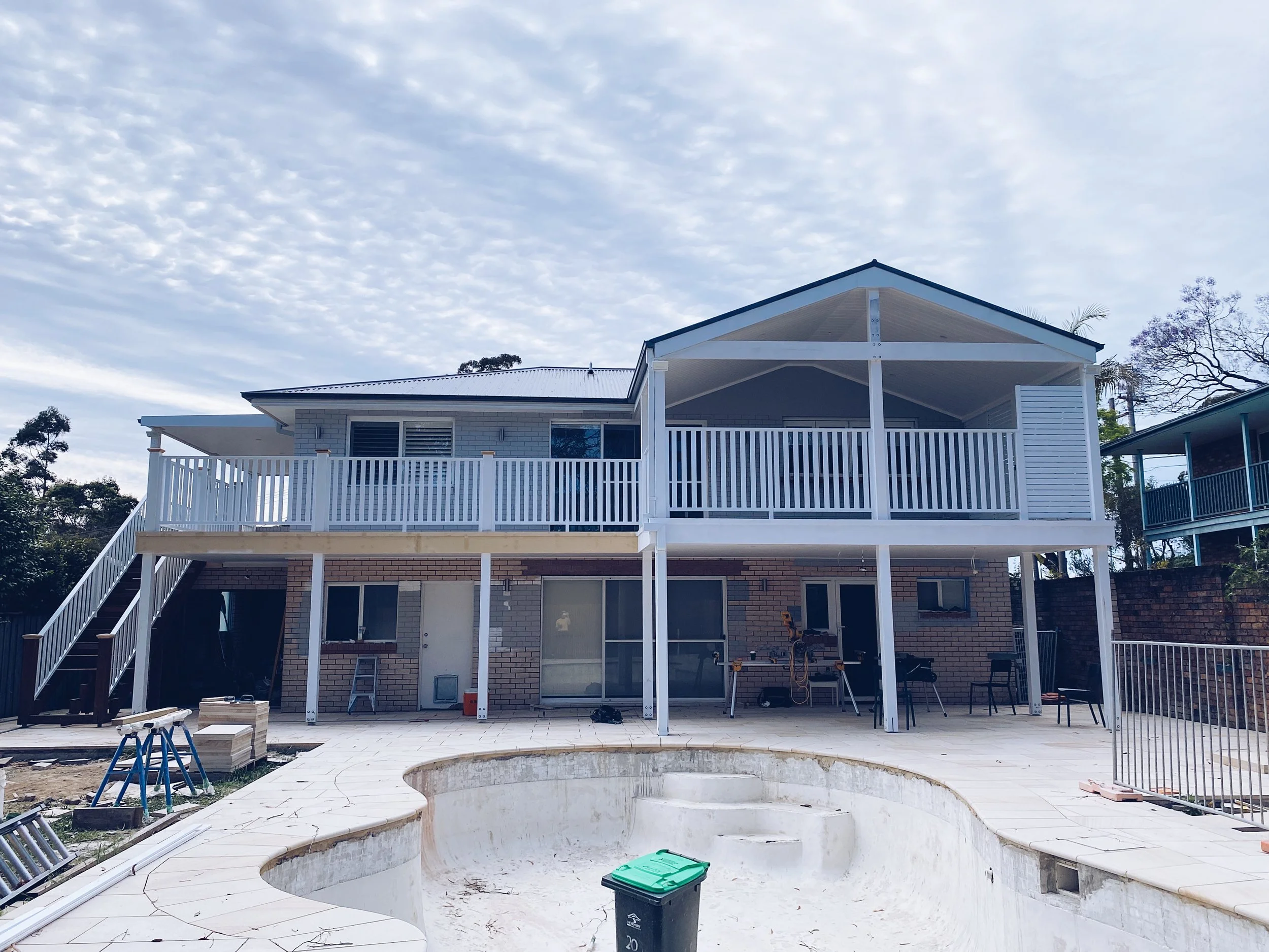 A two-story house with a white wooden balcony, under construction or renovation. There is a circular empty swimming pool in the backyard, construction tools and materials around, and a pile of tiles or bricks.