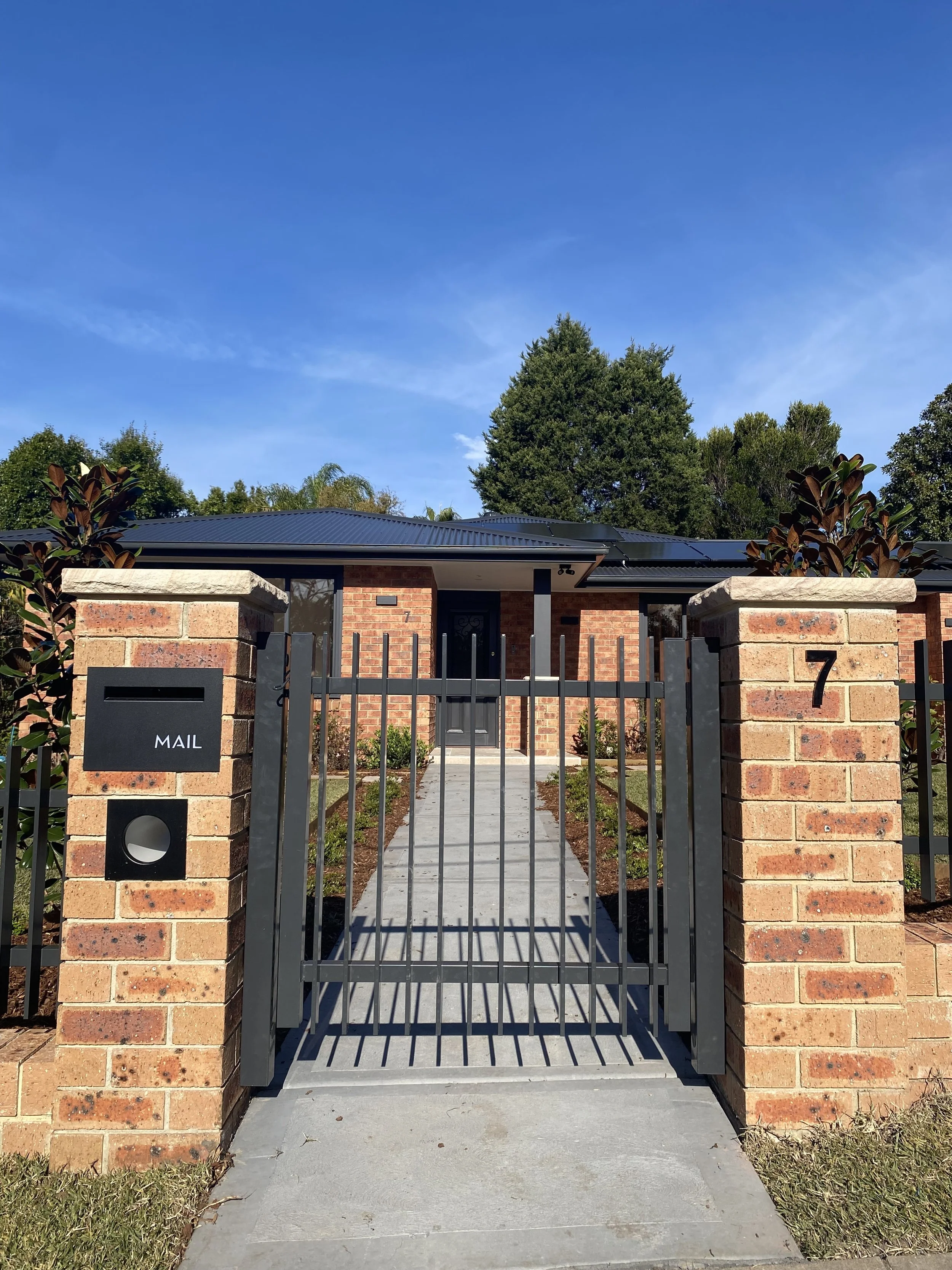Front view of a brick house with a gated entrance, mail slot, and house number 7, with a sidewalk leading to the front door, greenery on sides, and a large blue sky overhead.
