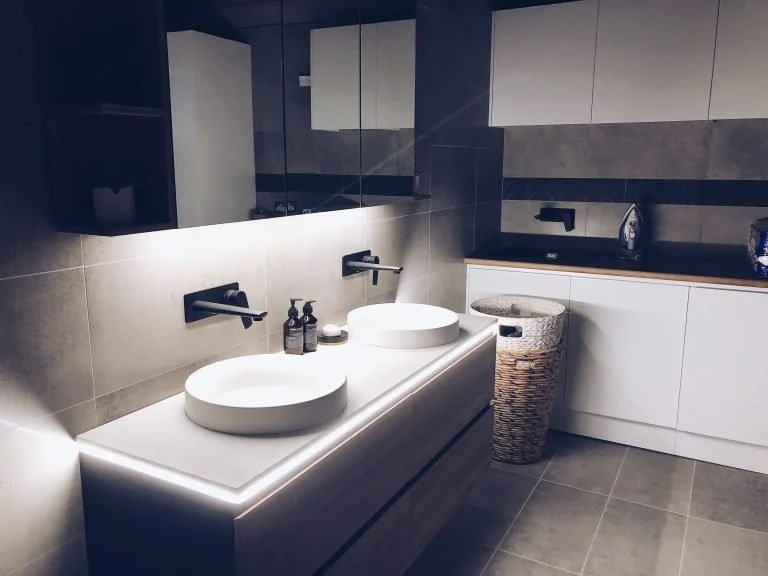 Modern bathroom with dual vessel sinks, large mirror, gray tiled wall, and white cabinetry.