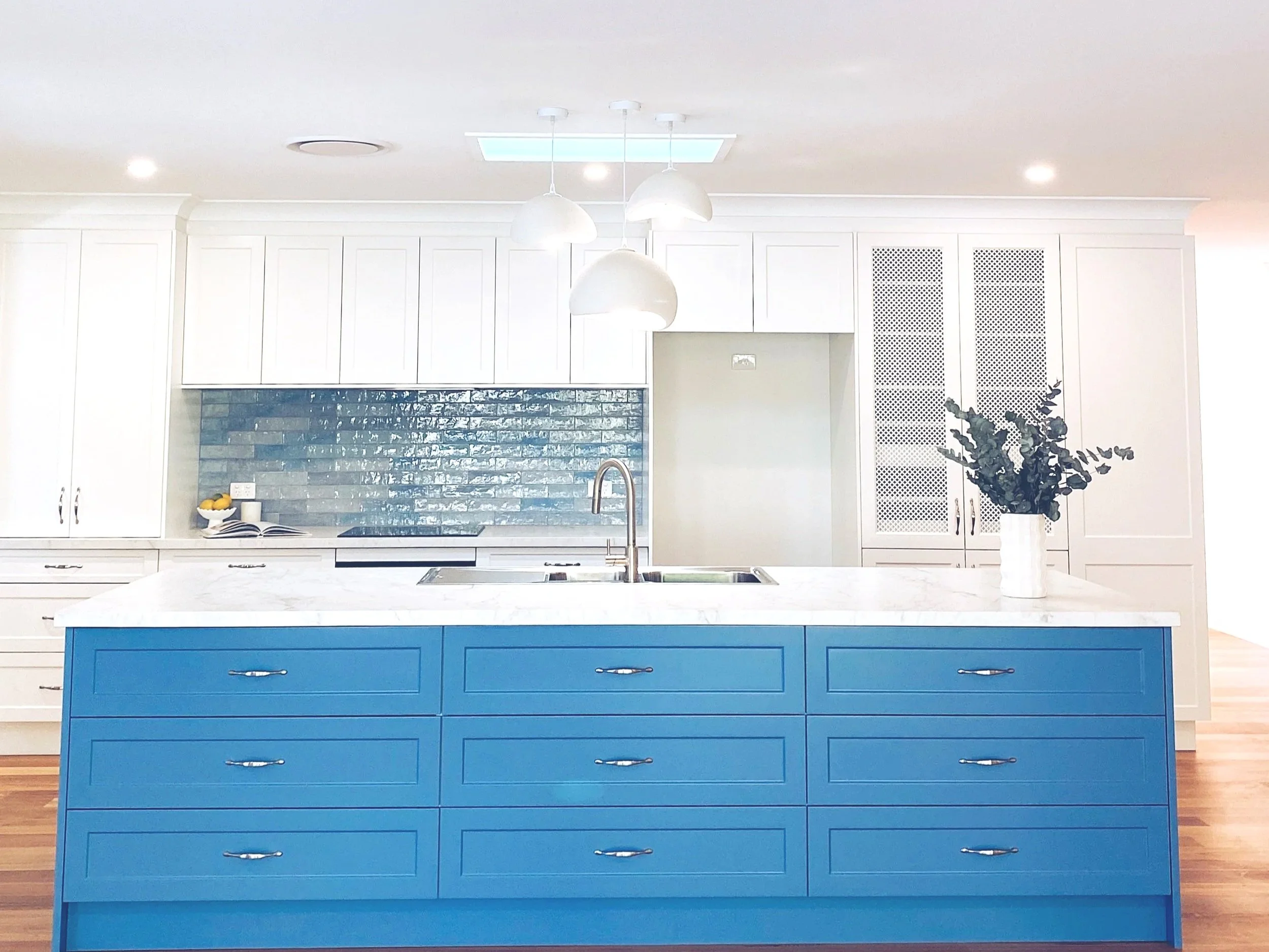 A modern kitchen with white cabinetry, a grey brick backsplash, and a blue kitchen island with multiple drawers and silver handles. A white vase with greenery decorates the island, and three white pendant lights hang above it. There is a window in the ceiling providing natural light.