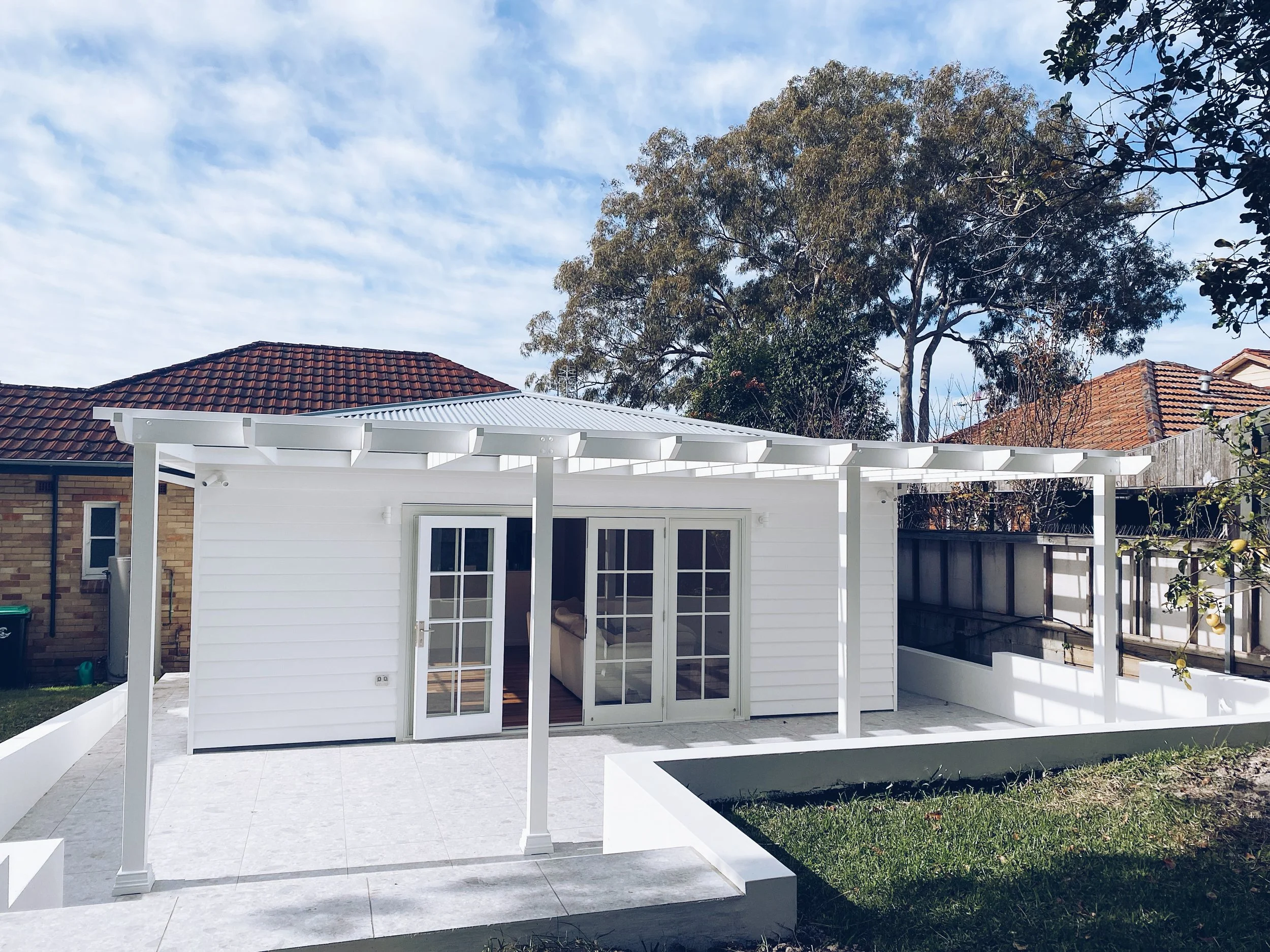 Backyard patio with white pergola, sliding glass door, tiled floor, lush green grass, and neighboring brick and tile-roofed houses under a partly cloudy sky.