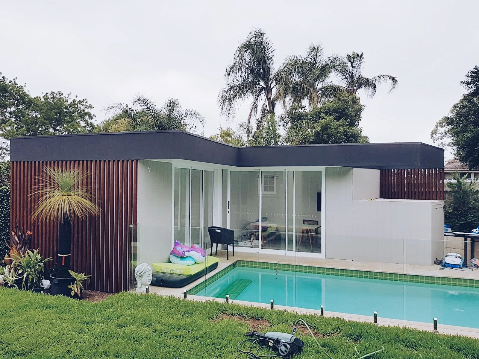 Modern backyard with a swimming pool, glass doors leading into a house, surrounded by green grass, tropical trees, and outdoor decorations.