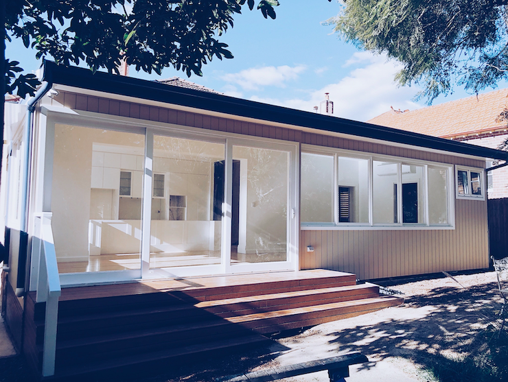 A modern house with large glass sliding doors, a set of wooden steps leading to the entrance, and a backyard shaded by trees.
