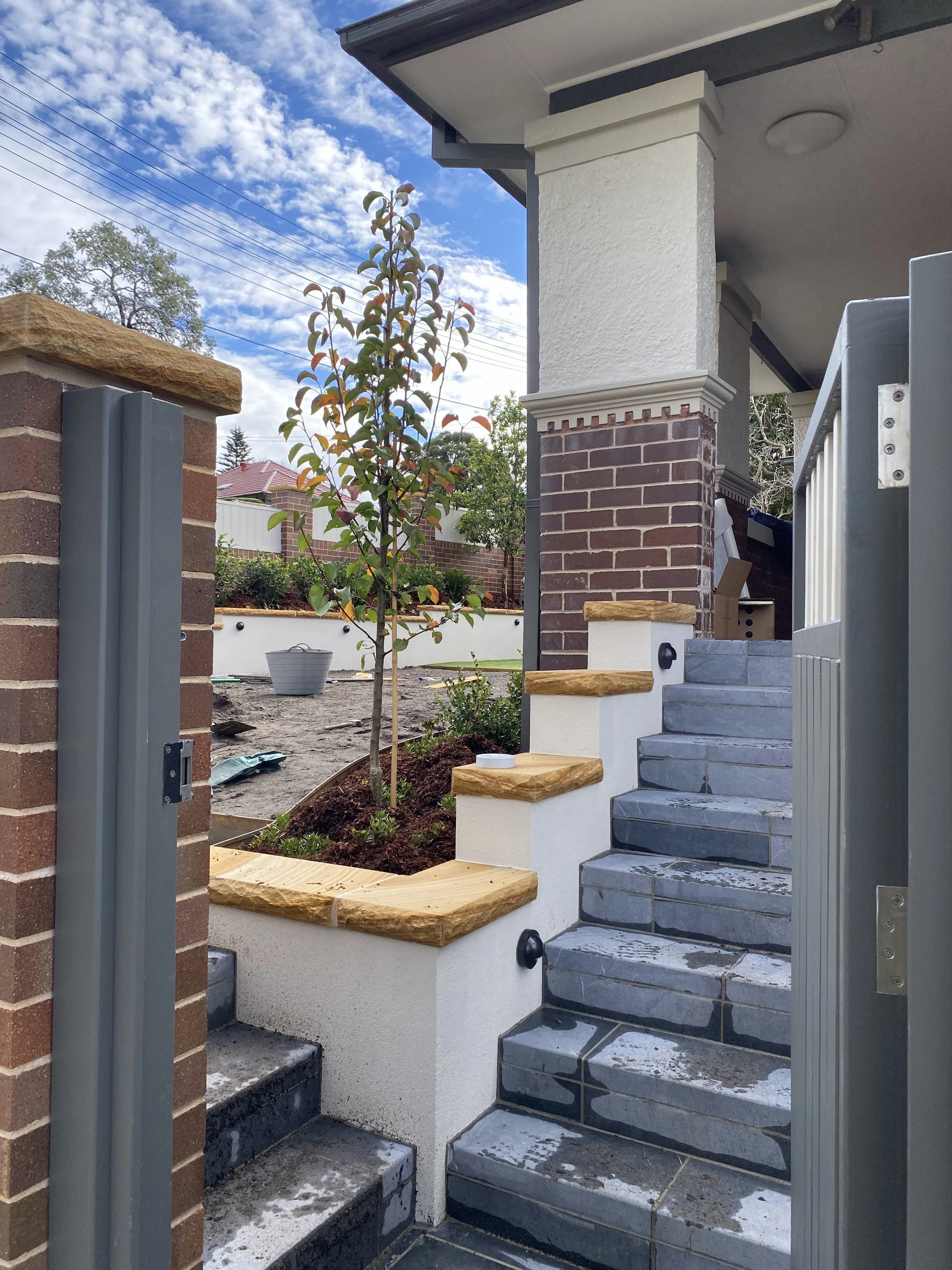 The image shows a newly constructed stairway leading to a house, with gray tiled steps, wooden accents on the edge of the white wall, and a small tree in a garden bed. There is a brick wall on the left and a metal gate at the entrance, with a clear s
