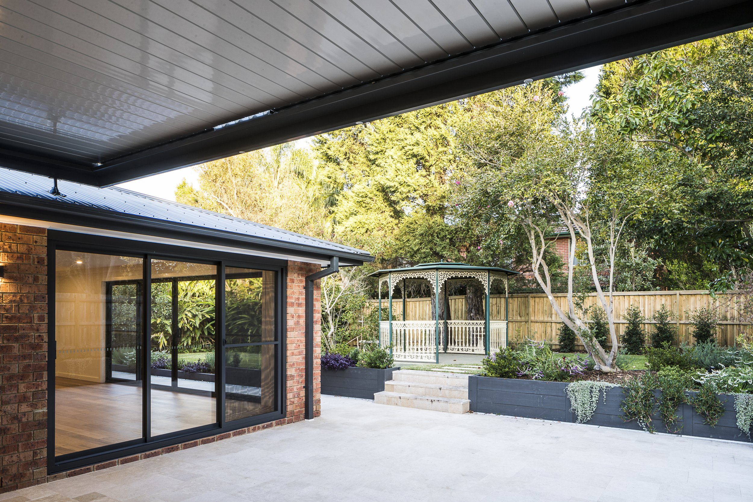 A backyard with a brick house, large sliding glass doors, a covered patio, a white wooden gazebo, and various green trees and plants.