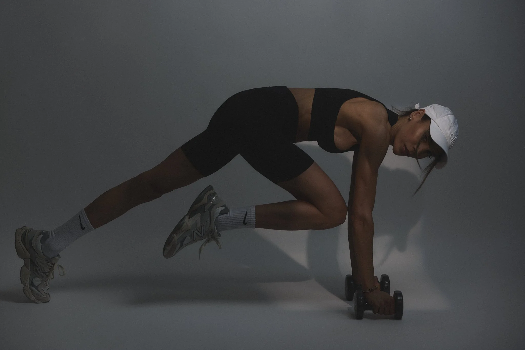 A woman in athletic wear, including a white cap, black sports bra, black shorts, and running shoes, is in a plank position on her hands, holding a dumbbell in one hand and stretching with a serious expression against a plain, gray background.