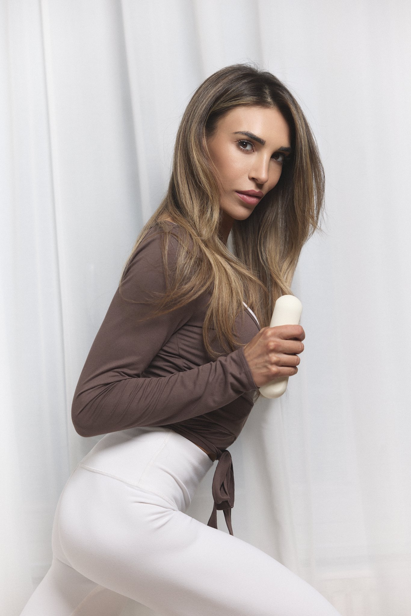 A woman doing Pilates, holding a white dumbbell set, wearing a brown top and white pants.