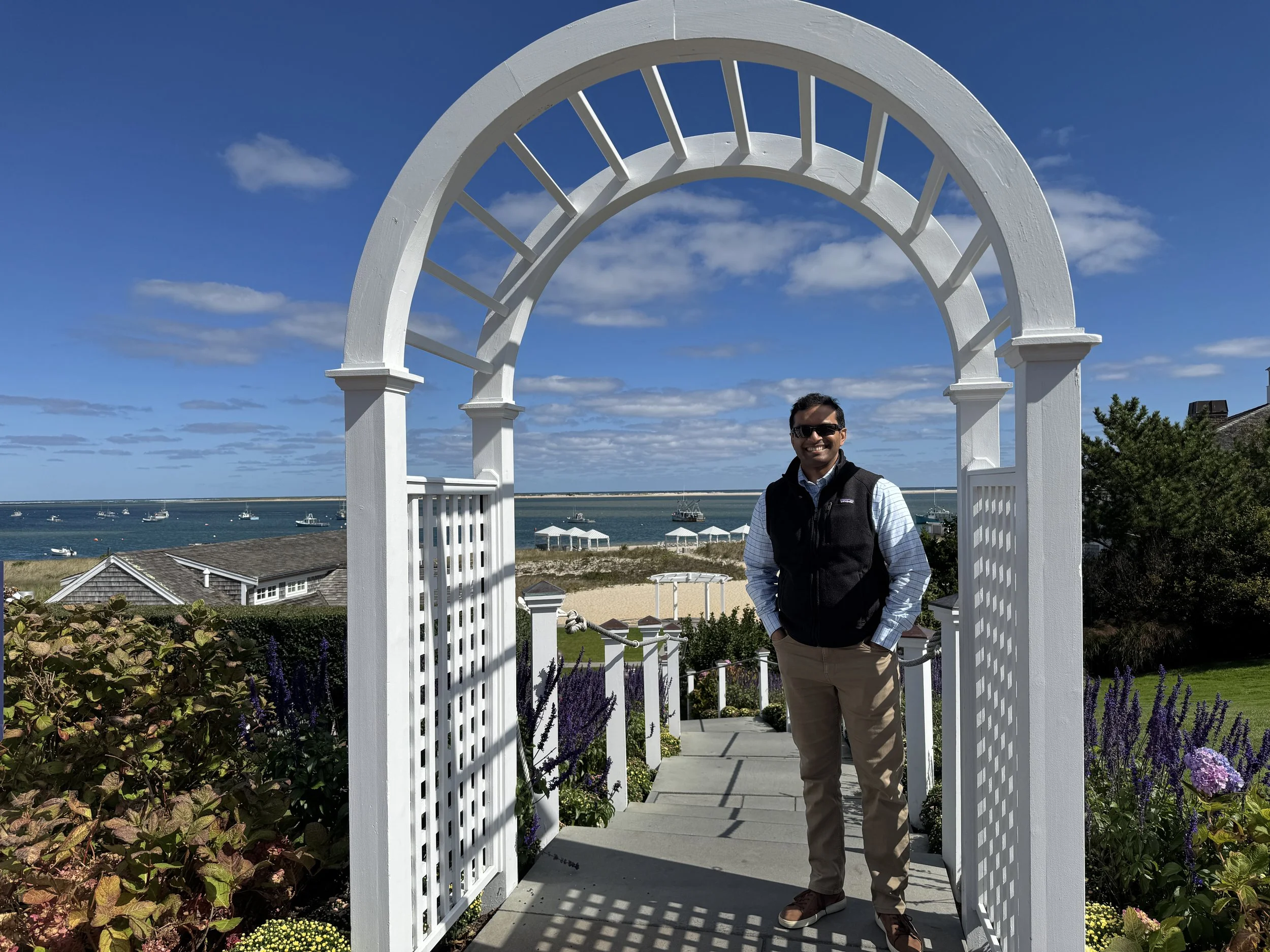 A man smiling and standing under a white wooden archway on a coastal background with boats, houses, and a blue sky with clouds.