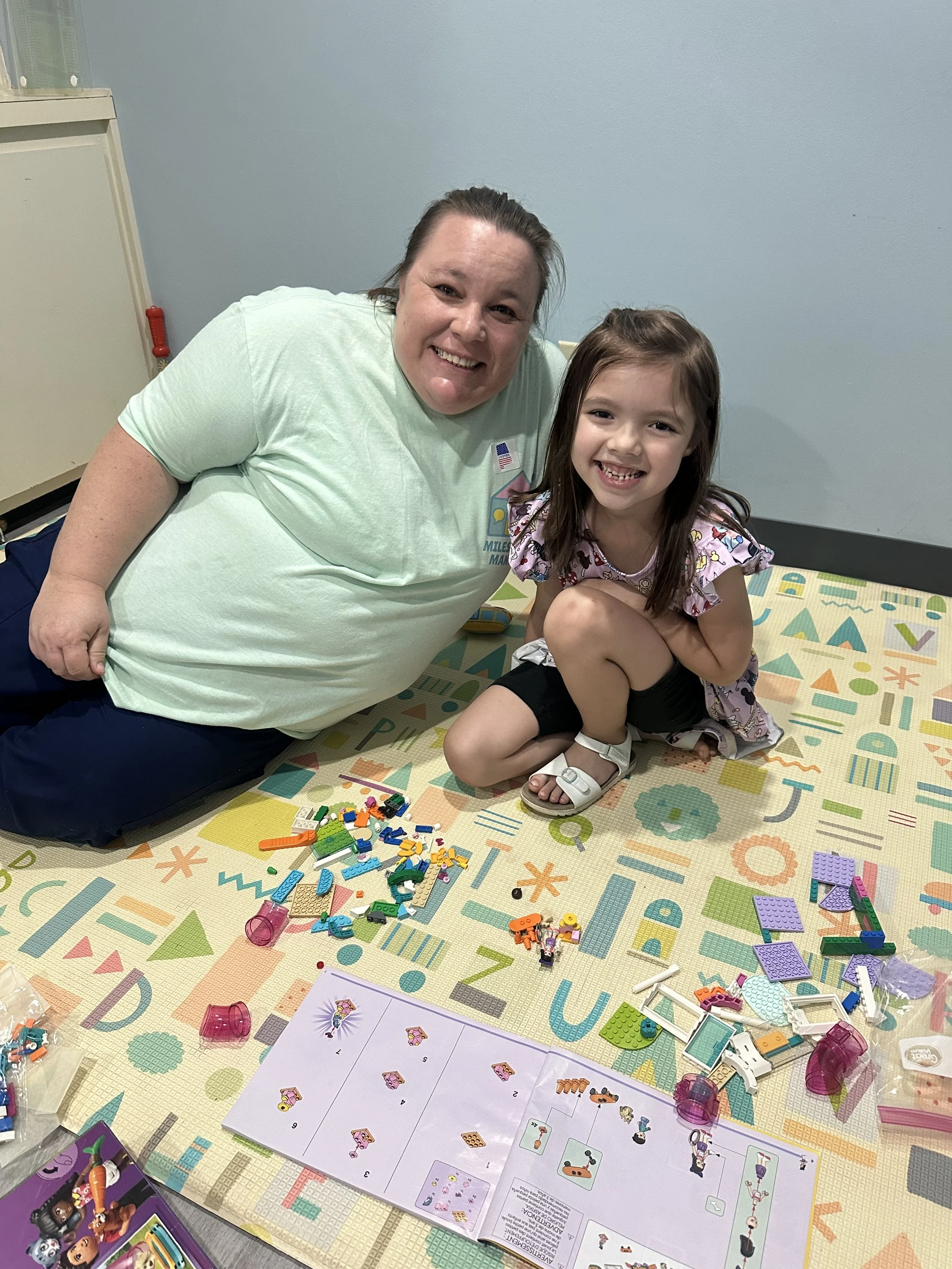 A woman and a girl sitting on a colorful play mat, smiling, surrounded by Lego pieces and instruction sheets.