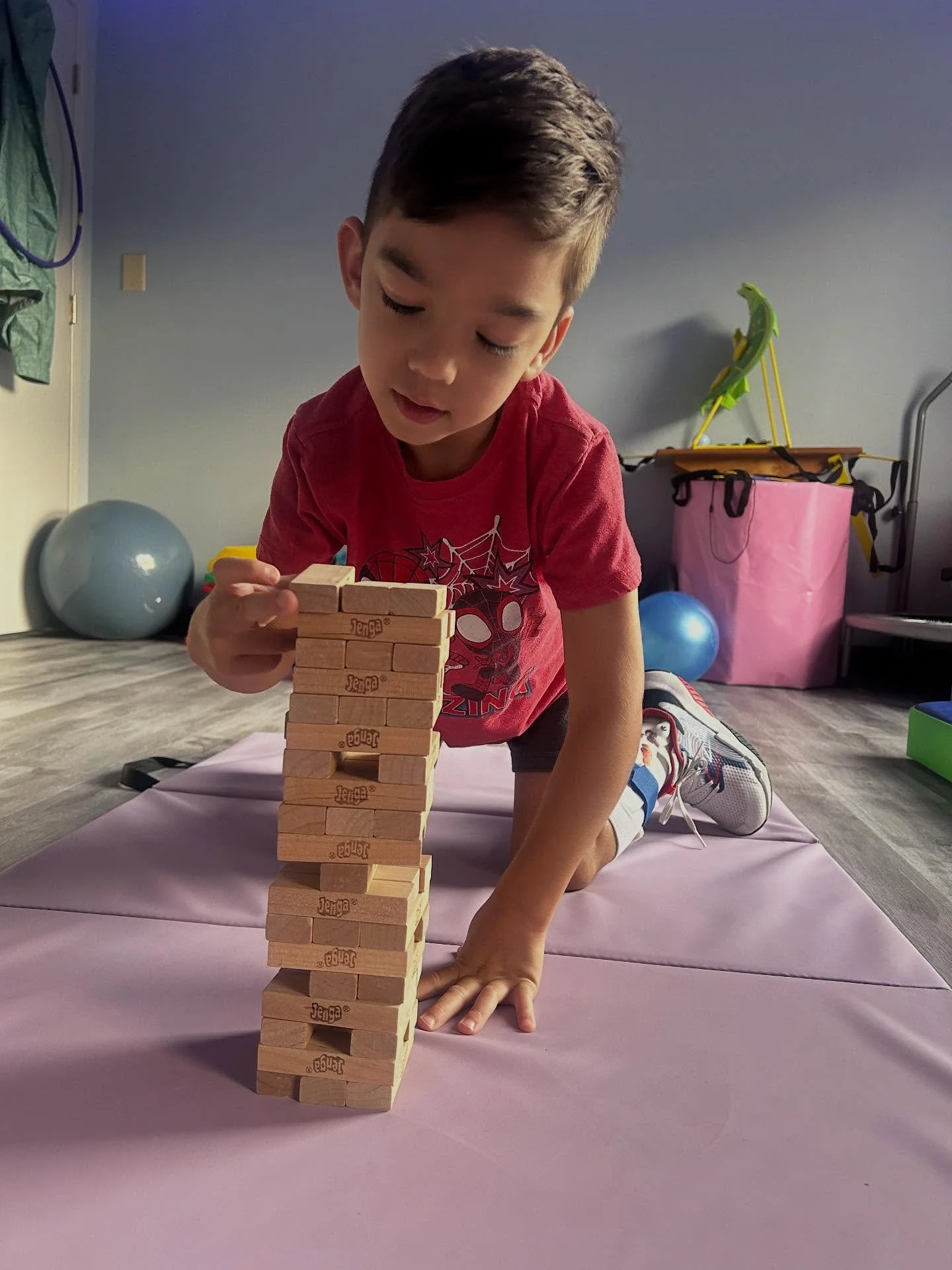 Young boy playing Jenga on a pink mat in a room with exercise equipment and a pink storage box.